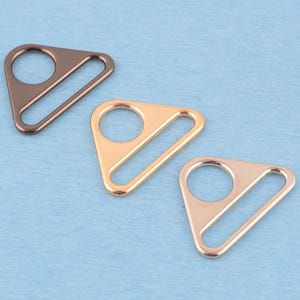 Op de afbeelding: Drie metalen riemverstellers in een driehoekige vorm met een ronde opening en een rechthoekige gleuf. De set bevat een donkerbrons, een goudkleurige en een roségouden afwerking, elk met een glad, gepolijst oppervlak en afgeronde randen.