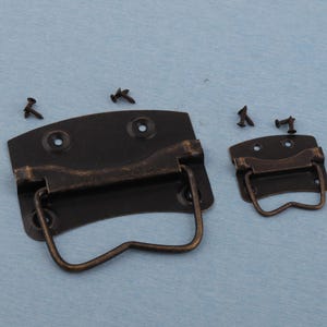 May include: Two antique bronze-colored metal trunk handles with matching screws. The handles have a curved top and a hinged, curved handle. The smaller handle is next to the larger one, with screws scattered around.