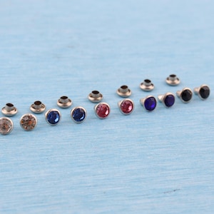 Może przedstawiać: Kolekcja metalowych nitów z kolorowymi, okrągłymi kamieniami szlachetnymi. Kamienie szlachetne są w różnych kolorach, w tym przezroczystym, brązowym, niebieskim, różowym i czarnym. Nity są ułożone na jasnoniebieskiej powierzchni.