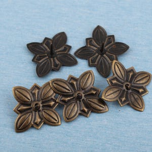 以下が含まれることがあります： ブロンズ色の花型装飾画鋲が5つ。各画鋲は、中央の穴と取り付け用のピンを備えた、詳細な花の模様が特徴です。画鋲は水色の表面に配置されています。