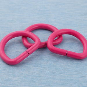 Pu&ograve; includere: Tre anelli metallici rosa brillante a forma di D sono interconnessi su uno sfondo azzurro. Gli anelli hanno una finitura liscia e lucida e un piccolo spazio su un lato. La disposizione suggerisce un focus sull'artigianato o sulla gioielleria.