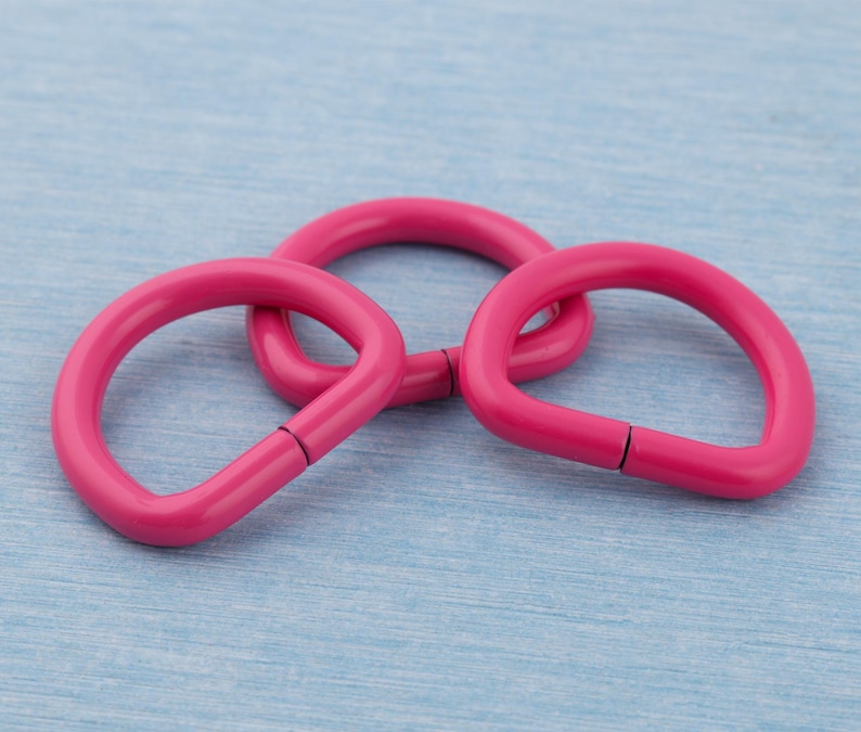 Pu&ograve; includere: Tre anelli metallici rosa brillante a forma di D sono interconnessi su uno sfondo azzurro. Gli anelli hanno una finitura liscia e lucida e un piccolo spazio su un lato. La disposizione suggerisce un focus sull'artigianato o sulla gioielleria.