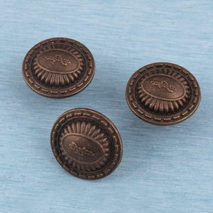 Pode incluir: Três puxadores de armário em bronze antigo com um design oval central e uma borda externa decorativa. Os puxadores estão dispostos sobre uma superfície azul clara. Os puxadores são provavelmente usados para móveis ou fins decorativos.