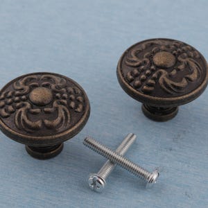 May include: Two antique bronze cabinet knobs with decorative floral patterns and a central circular design. Two silver screws are included. These knobs are ideal for adding a vintage touch to furniture.