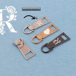 May include: Four metal zipper pulls in silver, gold, and black, with a blue background. One gold pull has a white heart, and two have a floral design with the letter "R". The text "your logo" is also visible.