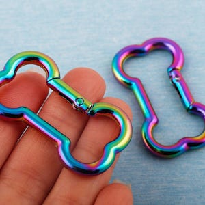 May include: Two iridescent rainbow-colored metal lobster clasps. The clasps have a unique clover-like shape and a spring-loaded gate for secure closure. The clasps are held in a hand against a light blue background.