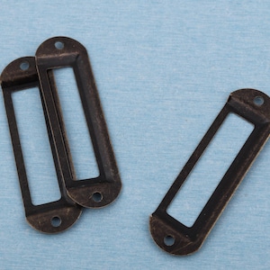 May include: Three antique bronze metal label holders. Each has a rectangular opening and two small holes for mounting. The label holders have a distressed finish, giving them a vintage appearance.