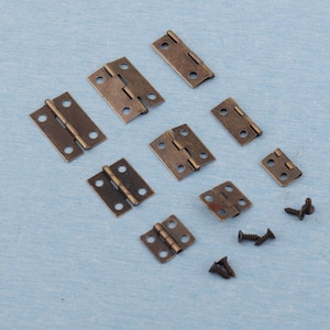 Könnte beinhalten: Eine Sammlung kleiner, antik bronzefarbener Scharniere und Schrauben. Die Scharniere sind in verschiedenen Größen und Formen erhältlich und verfügen über mehrere Schraubenlöcher. Das Zubehör ist auf einer hellblauen Oberfläche angeordnet, ideal für kleine Holzbearbeitungsprojekte.