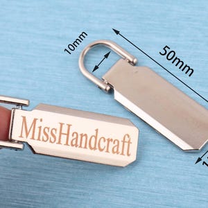 Peut inclure: Deux tirettes de fermeture &eacute;clair en m&eacute;tal argent&eacute; de forme rectangulaire avec une boucle sup&eacute;rieure arrondie. L'une des tirettes porte le texte grav&eacute; "MissHandcraft". Les dimensions indiqu&eacute;es sont 50mm de long, 15mm de large et 10mm pour l'ouverture de la boucle.