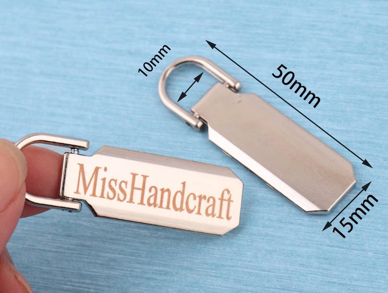 Peut inclure: Deux tirettes de fermeture &eacute;clair en m&eacute;tal argent&eacute; de forme rectangulaire avec une boucle sup&eacute;rieure arrondie. L'une des tirettes porte le texte grav&eacute; "MissHandcraft". Les dimensions indiqu&eacute;es sont 50mm de long, 15mm de large et 10mm pour l'ouverture de la boucle.