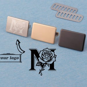 Puede incluir: Tres piezas rectangulares de metal en plata, oro y negro, con una rosa y la letra "M" grabadas en una de ellas. También hay dos piezas ranuradas de metal plateado. También se muestra un gráfico en blanco y negro de una rosa y la letra "M".