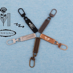 Könnte beinhalten: Eine Sammlung von Metall-Reißverschlussanhängern in verschiedenen Farben, darunter Schwarz, Silber und Bronze, die sternförmig angeordnet sind. Einige Anhänger weisen dekorative Designs auf. Der Hintergrund ist eine hellblaue Oberfläche.