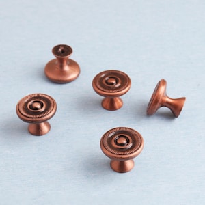 Könnte beinhalten: Fünf kupferfarbene Schrankknöpfe mit kreisförmigem, geriffeltem Design. Die Knöpfe haben einen Stiel und eine abgerundete Oberseite und sind auf einer hellblauen Oberfläche angeordnet. Diese dekorativen Knöpfe sind für Möbel geeignet.