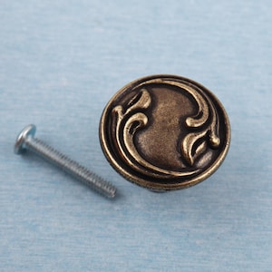 Könnte beinhalten: Ein runder Schrankknauf in antik-messingfarbenem Finish mit einem verzierten Wirbeldesign. Eine silberne Schraube befindet sich links vom Knauf. Der Knauf ist wahrscheinlich für Möbel oder dekorative Zwecke bestimmt.