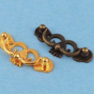 May include: Two metal drawer pulls, one with a polished gold finish and one with an antique bronze finish. Each pull features a decorative backplate with a central fan-like detail and a curved, hinged handle attached to two circular mounting points.