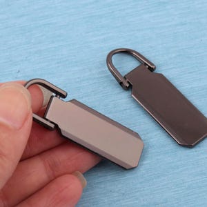 Peut inclure: Deux tirettes de fermeture &eacute;clair rectangulaires en m&eacute;tal avec une finition gris acier fonc&eacute; lisse. Chaque tirette pr&eacute;sente une boucle sup&eacute;rieure arrondie pour la fixation et un corps plat et allong&eacute; aux bords biseaut&eacute;s.