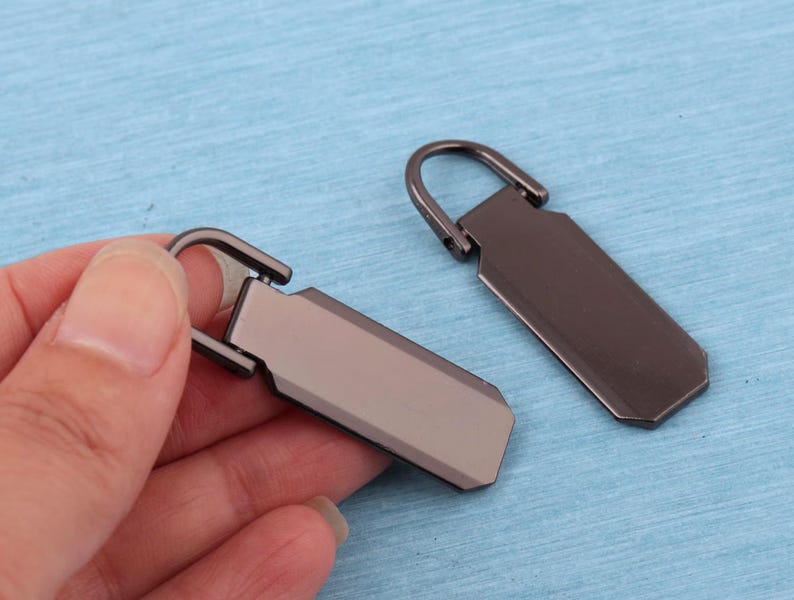 Peut inclure: Deux tirettes de fermeture &eacute;clair rectangulaires en m&eacute;tal avec une finition gris acier fonc&eacute; lisse. Chaque tirette pr&eacute;sente une boucle sup&eacute;rieure arrondie pour la fixation et un corps plat et allong&eacute; aux bords biseaut&eacute;s.