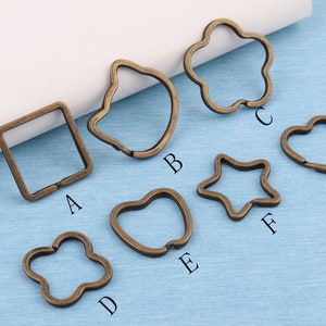 May include: Assortment of eight different metal keyrings in various shapes, including a square, heart, star, flower, and more. The keyrings are all a bronze colour.