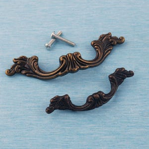 May include: Two ornate, curved drawer pulls made of dark bronze-toned metal with intricate scrollwork and leaf-like detailing. The hardware set includes two small silver-colored metal screws for installation, positioned above the larger handle.