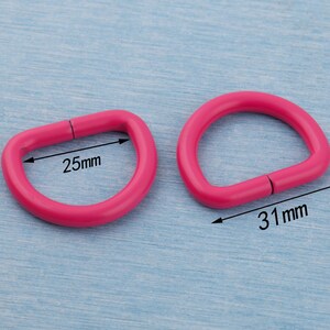 Pu&ograve; includere: Due anelli a D rosa acceso. Uno misura 25 mm di larghezza, l'altro 31 mm. Questi anelli metallici sono utilizzati in vari lavori artigianali e accessori. Gli anelli a D sono mostrati su una superficie azzurra.