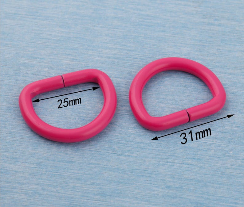 Pu&ograve; includere: Due anelli a D rosa acceso. Uno misura 25 mm di larghezza, l'altro 31 mm. Questi anelli metallici sono utilizzati in vari lavori artigianali e accessori. Gli anelli a D sono mostrati su una superficie azzurra.