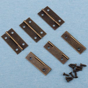 Op de afbeelding: Zes kleine antieke messing scharnieren en bijpassende schroeven zijn gerangschikt op een lichtblauwe ondergrond. Elk scharnier is rechthoekig met afgeronde hoeken en drie schroefgaten aan elke kant. De scharnieren zijn verbonden door een centrale pen.