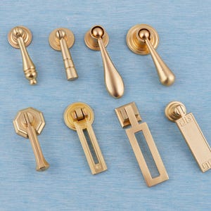 以下が含まれることがあります： さまざまなデザインの、8つの金色のキャビネットプル。ティアドロップ型、円筒型、長方形の形状があり、すべて金色の仕上げです。明るい青色の表面に配置されています。