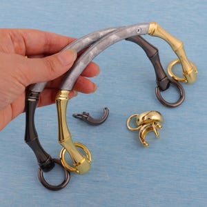 Può includere: Due manici per borse con finitura argento e nera e dettagli dorati. I manici hanno un design curvo e sono in metallo. Sono inclusi anche due fermagli dorati aggiuntivi.