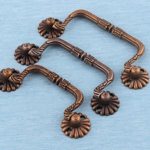 May include: Three antique bronze-colored metal drawer pulls featuring a curved handle design with textured detailing and circular mounting plates that have a scalloped edge pattern. Each pull is secured by two round bases with screw holes.