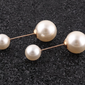 Op de afbeelding: Twee broches van goudkleur met witte parel accenten. Elke broche heeft twee parels, een groter dan de andere.
