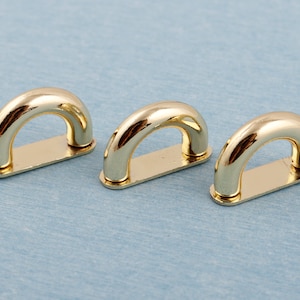 Op de afbeelding: Drie gouden metalen D-ringen, elk met een glad, afgerond oppervlak. De ringen zijn in een rij gerangschikt, met de linker ring iets dichter bij de kijker.