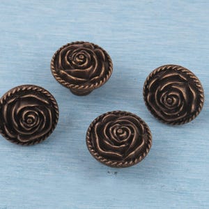 May include: Four bronze-colored cabinet knobs with a rose design. Each knob features a detailed rose in the center, surrounded by a rope-like border. The knobs are round and have a vintage aesthetic, suitable for furniture restoration or decorative projects.