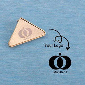 May include: A gold-colored, triangular pin with rounded edges. The pin features a pumpkin-like design and the text "Monster.T". The background is a textured, light blue surface. A logo with the text "Your Logo" is also present.