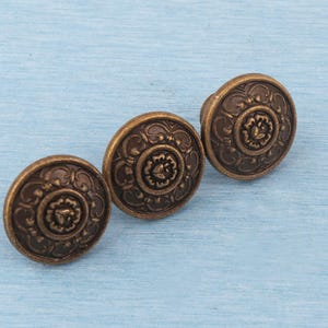 May include: Three antique bronze cabinet knobs with a floral design. The knobs are round with a raised center and decorative detailing around the edges. They are arranged on a light blue surface.