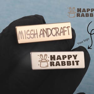 May include: Two rectangular metal pins. The top pin has the text "MISSH AND CRAFT" engraved. The bottom pin has the text "HAPPY RABBIT" with a rabbit in a hat logo. The Happy Rabbit logo is also displayed above the pins.