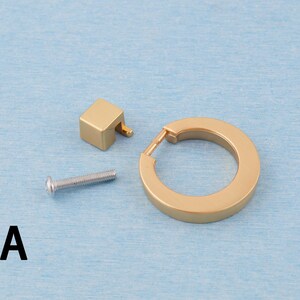 Op de afbeelding: Een set goudkleurige kastbeslagcomponenten, waaronder een vierkante knop, een cirkelvormige handgreep en een zilveren schroef. De hardware is ontworpen voor meubels en heeft een moderne, minimalistische uitstraling. De letter "A" is zwart gedrukt.