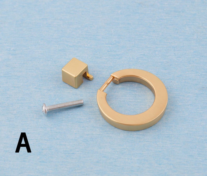 Op de afbeelding: Een set goudkleurige kastbeslagcomponenten, waaronder een vierkante knop, een cirkelvormige handgreep en een zilveren schroef. De hardware is ontworpen voor meubels en heeft een moderne, minimalistische uitstraling. De letter "A" is zwart gedrukt.