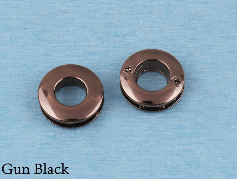 May include: Two circular metal grommets with a polished gun black finish. The grommet on the right features two small visible screws on its surface, while the grommet on the left is smooth. The text Gun Black is printed in the bottom left corner.