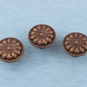 Puede incluir: Tres tiradores de armario de color bronce con diseño floral. Cada tirador presenta una flor central, detalles en forma de pétalos y un borde tipo cuerda. Los tiradores están sobre un fondo azul claro.
