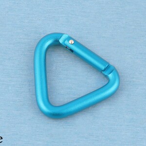 May include: A triangular carabiner in a bright blue metallic finish. The carabiner features a spring-loaded gate mechanism with a small silver-colored metal pin hinge. The overall shape is a rounded triangle with a smooth, matte-like texture.