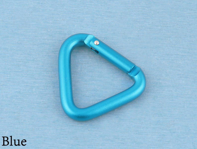 May include: A triangular carabiner in a bright blue metallic finish. The carabiner features a spring-loaded gate mechanism with a small silver-colored metal pin hinge. The overall shape is a rounded triangle with a smooth, matte-like texture.