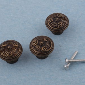 Può includere: Tre pomelli per mobili in bronzo antico con disegni ornati e a spirale. I pomelli sono rotondi con un motivo centrale in rilievo e una finitura scura e invecchiata. Sono visibili anche due viti argentate, che suggeriscono l'hardware di installazione.