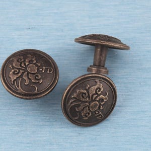 Pode incluir: Dois puxadores de armário antigos, cor bronze, com desenhos florais. Um puxador está solto, mostrando a haste e a parte de trás do puxador. Os puxadores têm um padrão floral em relevo detalhado sobre um fundo azul claro.