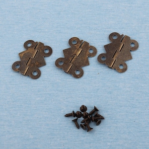 Könnte beinhalten: Drei antik-bronze farbene Scharniere mit dekorativen Kanten und mehreren Schraubenlöchern sind auf einer hellblauen Oberfläche angeordnet. Ein kleiner Haufen passender bronzefarbener Schrauben ist ebenfalls vorhanden, was auf ein Set für ein kleines Holzbearbeitungs- oder Bastelprojekt hindeutet.