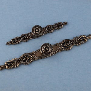 以下が含まれることがあります： 花柄とスクロールワークのディテールが施された、2つのアンティークブロンズキャビネットプル。各プルは、中央に花の模様の円形メダリオンがあり、長さは約15cmです。