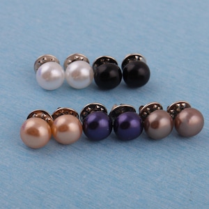 Puede incluir: Un conjunto de pendientes de botón con aspecto de perla en varios colores, incluyendo blanco, negro, melocotón, morado y gris. Cada pendiente tiene una cuenta redonda y brillante unida a una base de metal plateado. Sobre una superficie azul claro.