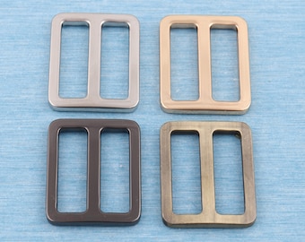 May include: Four metal slider buckles in different finishes: silver, gold, black, and antique brass. Each buckle has two parallel slots.