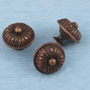 May include: Three bronze-coloured cabinet knobs with a floral design. The knobs have a rounded shape with a central sphere and detailed, curved lines radiating outwards. The knobs are on a light blue surface.