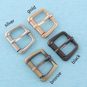 May include: Four metal belt buckles in different finishes: gold, silver, bronze, and black. Each buckle has a rectangular shape with a smooth, polished surface.