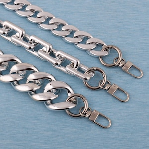 Puede incluir: Tres correas de cadena de metal plateado con cierres de mosquetón. Las cadenas tienen diferentes tamaños de eslabones, con un diseño grueso y redondeado. El fondo es una superficie azul claro.